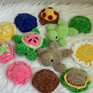 May include: A collection of small, plush, crocheted items in various shapes and colors, including a watermelon turtle, a plain green turtle, a brown turtle, a yellow flower, a pink strawberry, a yellow lemon slice, and several round textured discs.