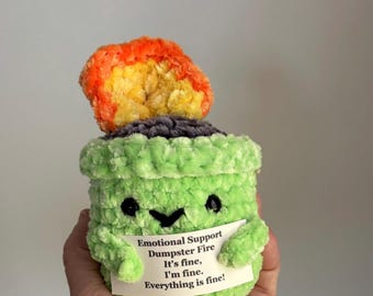 May include: A handmade, green crochet plushie shaped like a pot with a smiling face and arms holding a sign. The sign reads "Emotional Support Dumpster Fire. It's fine, I'm fine. Everything is fine!" A flame made of orange, yellow, and red yarn sits atop the pot.