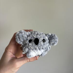 Könnte beinhalten: Ein kleines, graues und weißes gehäkeltes Koala-Plüschtier mit schwarzen Augen. Der Koala wird in der Hand einer Person gehalten.