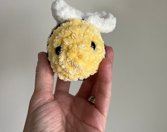May include: A crocheted bee plush toy with yellow and white yarn. The bee has black eyes and a white fuzzy body.