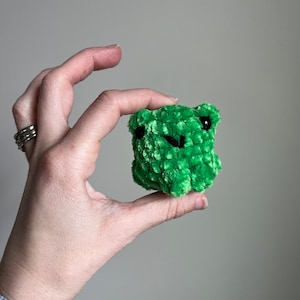 May include: A small green crocheted frog toy with black eyes. The frog is being held in a hand.
