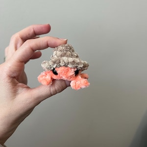 May include: A handmade, plush crab in shades of coral and beige. The crab is wearing a small, textured hat. The crab's eyes are black. The plush toy is held in a hand.
