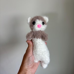 Puede incluir: Un peluche de hurón hecho a mano con cuerpo blanco, cabeza marrón y blanca y nariz rosa. El hurón tiene ojos negros y está hecho de material suave y esponjoso. Un juguete de peluche lindo y tierno.