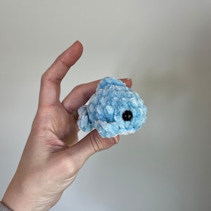 May include: A small, blue crocheted fish toy with a black eye. The fish is being held in a hand.