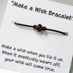 Peut inclure: Un bracelet de vœux en cuir marron avec un nœud, présenté sur une carte blanche. La carte porte l'inscription "Make a Wish Bracelet" et des instructions.