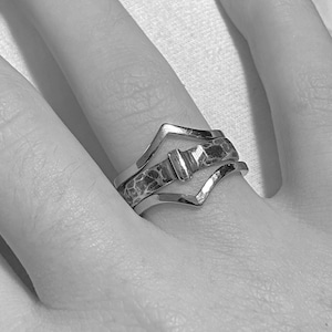 May include: A silver ring set with three bands. The top band has a V-shape, the middle band has a hammered texture, and the bottom band has a V-shape. The ring is on a finger.