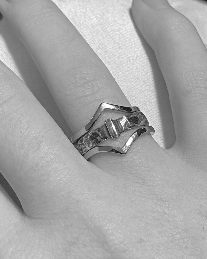 May include: A silver ring set with three bands. The top band has a V-shape, the middle band has a hammered texture, and the bottom band has a V-shape. The ring is on a finger.