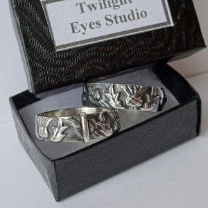 May include: Two silver rings with detailed floral and bird designs rest in a black box. The rings have a vintage, handcrafted appearance. The box has a textured exterior and a label that reads "Twilight Eyes Studio".