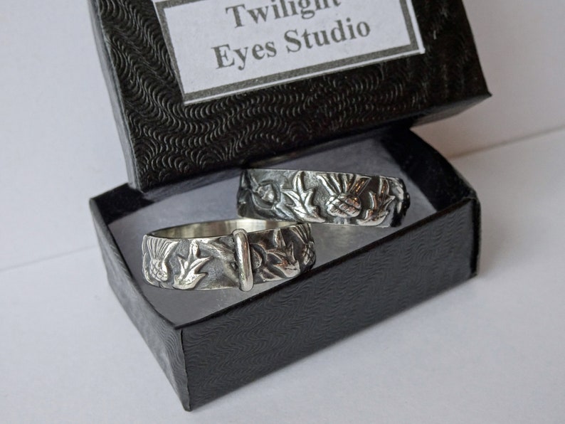 May include: Two silver rings with detailed floral and bird designs rest in a black box. The rings have a vintage, handcrafted appearance. The box has a textured exterior and a label that reads "Twilight Eyes Studio".