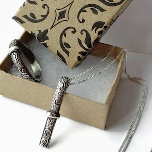 May include: A silver pendant necklace and ring set displayed in a decorative box. The jewelry features an ornate, engraved floral design. The pendant is a vertical bar shape, and the ring is a band. The box has a brown exterior with a black and white pattern.