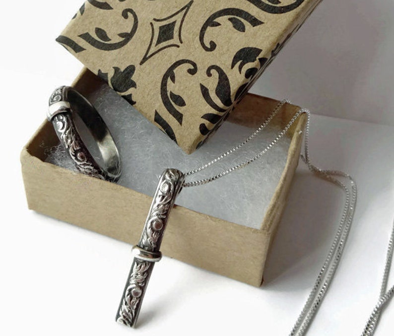 May include: A silver pendant necklace and ring set displayed in a decorative box. The jewelry features an ornate, engraved floral design. The pendant is a vertical bar shape, and the ring is a band. The box has a brown exterior with a black and white pattern.