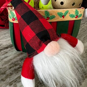 Pode incluir: Um gnomo festivo com uma longa barba branca, nariz rosa e chapéu e braços xadrez vermelhos e pretos. O gnomo está em frente a uma cesta decorativa com detalhes verdes, vermelhos e de madeira, e uma campainha dourada.