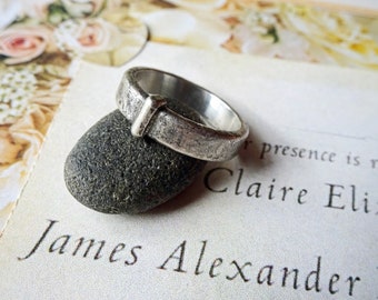 May include: A silver ring with a textured surface and a rectangular accent. The ring is resting on a dark gray stone, with a blurred background of a floral design and text that reads "James Alexander".