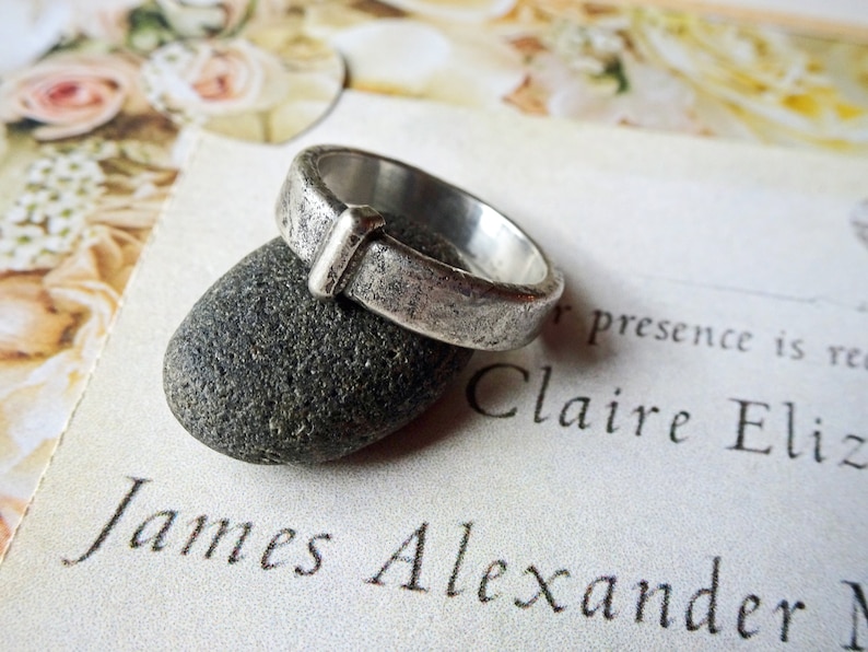 May include: A silver ring with a textured surface and a rectangular accent. The ring is resting on a dark gray stone, with a blurred background of a floral design and text that reads "James Alexander".