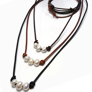 May include: Three necklaces with white pearls and leather cords in black and brown. Each necklace features a different number of pearls, with the longest necklace having five pearls. The cords are knotted to secure the pearls.