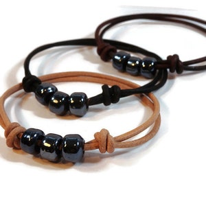 Può includere: Tre braccialetti con perline scure e lucenti e cordoncini in pelle. Un braccialetto è marrone chiaro, uno è nero e il terzo è marrone scuro. Ogni braccialetto ha una chiusura a nodo.