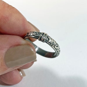 May include: A silver ring with an ornate, swirling floral design. The ring features a band with detailed scrollwork and a central section with two parallel, raised bands. The ring is held against a white background.
