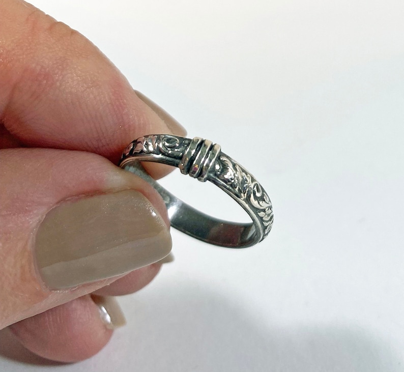 May include: A silver ring with an ornate, swirling floral design. The ring features a band with detailed scrollwork and a central section with two parallel, raised bands. The ring is held against a white background.