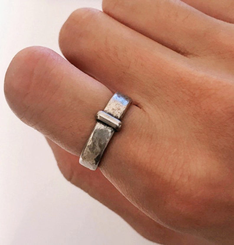 May include: A silver ring with a hammered texture and a rectangular bar design. The ring is worn on a finger, showcasing its simple yet elegant style. The ring's band is approximately 0.25 inches wide.