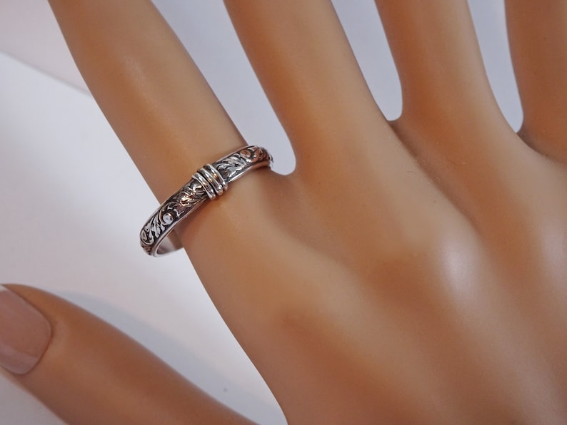 May include: A silver ring with an engraved floral pattern and a decorative band. The ring is on a finger, showcasing the intricate details of the design. The ring's band is approximately 0.25 inches wide.