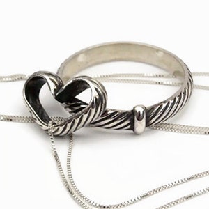 May include: A silver heart-shaped pendant with a textured design, connected to a matching silver chain. The pendant is intertwined with a silver band, creating a unique jewelry piece. The background is plain white.