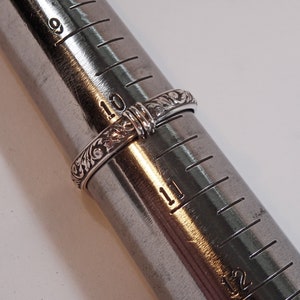 May include: A silver ring with an engraved floral pattern is displayed on a ring sizer. The ring is size 10, with the sizer showing measurements in inches. The ring's intricate design is visible.