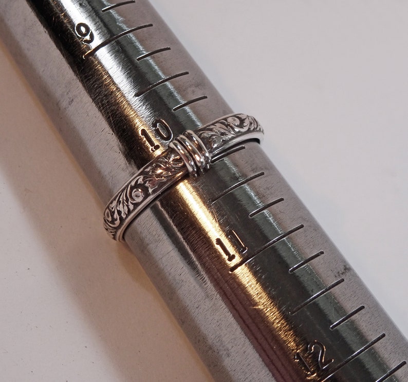 May include: A silver ring with an engraved floral pattern is displayed on a ring sizer. The ring is size 10, with the sizer showing measurements in inches. The ring's intricate design is visible.