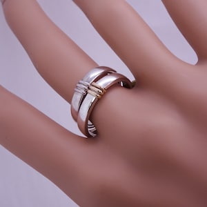 Könnte beinhalten: Ein silberner Ring mit goldenem Akzent wird auf einem Finger gezeigt. Der Ring hat ein einzigartiges Design mit zwei sich kreuzenden Bändern, die durch einen goldenen Balken verbunden sind. Ein Schmuckstück.
