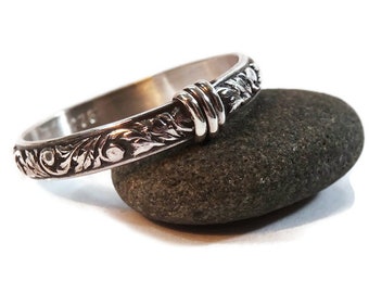 May include: A silver ring with an engraved floral pattern and a decorative knot design. The ring is resting on a dark gray stone, showcasing its intricate details and craftsmanship. The ring is a piece of jewelry.