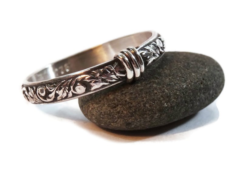 May include: A silver ring with an engraved floral pattern and a decorative knot design. The ring is resting on a dark gray stone, showcasing its intricate details and craftsmanship. The ring is a piece of jewelry.