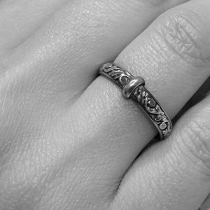 May include: A silver ring with an ornate floral design and a central knot-like detail. The ring is worn on a finger, showcasing its intricate carvings and polished finish. The ring's design is symmetrical, with detailed patterns.