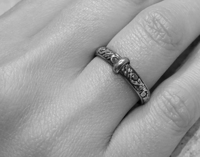May include: A silver ring with an ornate floral design and a central knot-like detail. The ring is worn on a finger, showcasing its intricate carvings and polished finish. The ring's design is symmetrical, with detailed patterns.