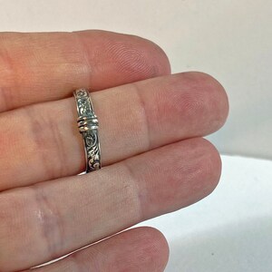 May include: A silver ring with an ornate floral design. The ring has a detailed pattern of swirling leaves and flowers. The band is approximately 0.1 inches wide. The ring is held between two fingers.