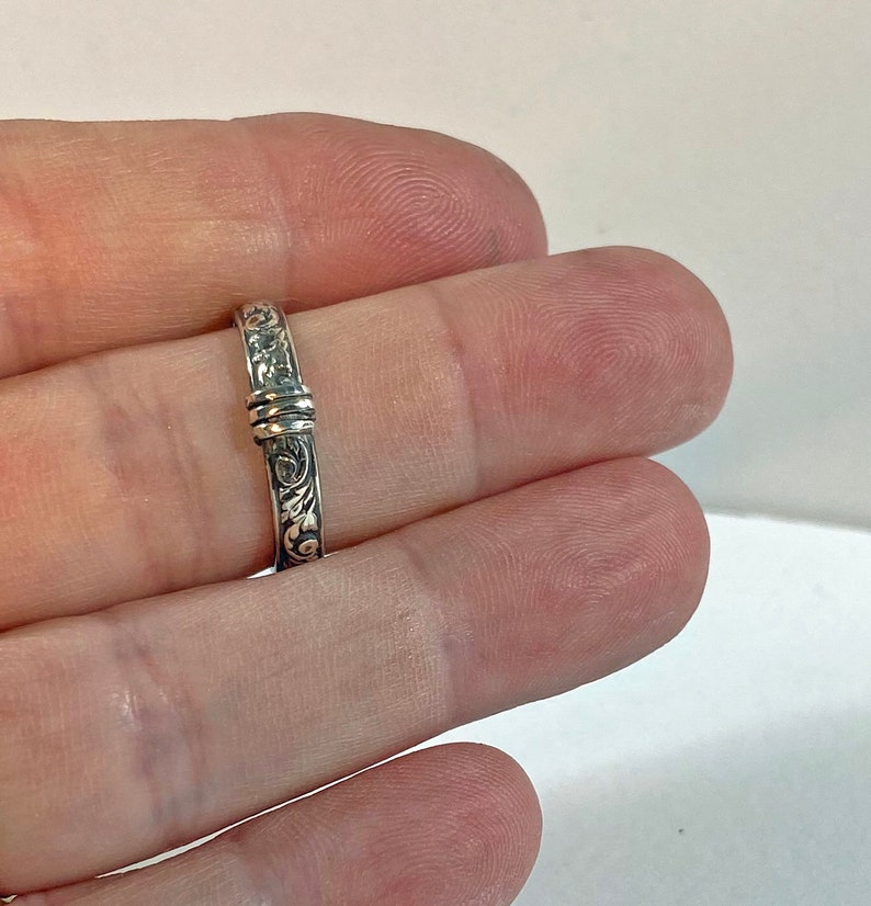 May include: A silver ring with an ornate floral design. The ring has a detailed pattern of swirling leaves and flowers. The band is approximately 0.1 inches wide. The ring is held between two fingers.