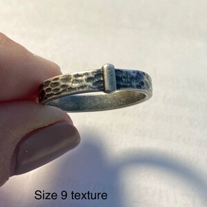 May include: A silver ring with a textured band and a rectangular accent. The ring is size 9. The band has a hammered texture, and the rectangular accent is smooth. The ring is held by a person.