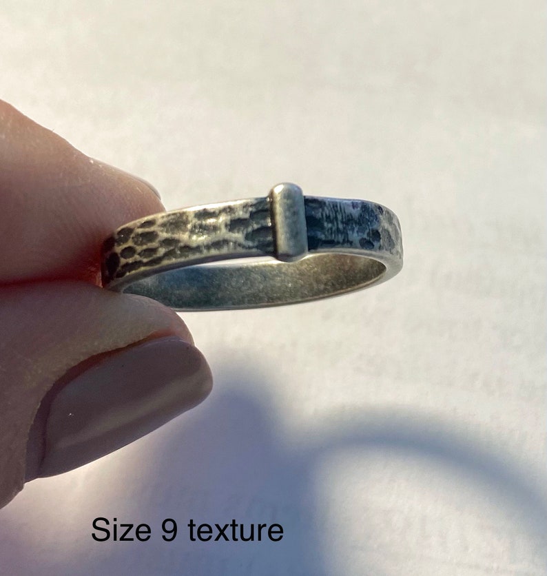 May include: A silver ring with a textured band and a rectangular accent. The ring is size 9. The band has a hammered texture, and the rectangular accent is smooth. The ring is held by a person.
