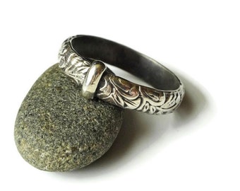 May include: A silver ring with an engraved floral pattern and a small silver clasp. The ring is resting on a gray stone against a white background. The ring's design features detailed, repeating floral motifs.