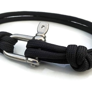 May include: Black rope bracelet with a stainless steel clasp. The bracelet features a nautical design with a knotted rope detail and a secure clasp. The bracelet is a stylish accessory.