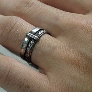May include: A silver ring with a buckle design. The ring has a detailed floral pattern on the band and a rectangular buckle element. The ring is worn on a finger, showcasing its intricate craftsmanship and vintage style.