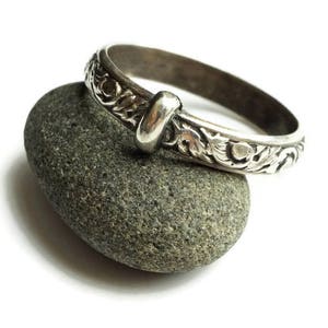 May include: A silver-colored ring with an ornate floral design and a knot detail. The ring is resting on a gray stone against a white background. The ring's intricate pattern is visible.