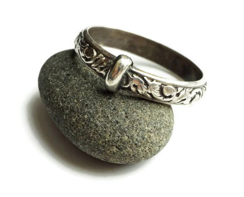 May include: A silver-colored ring with an ornate floral design and a knot detail. The ring is resting on a gray stone against a white background. The ring's intricate pattern is visible.