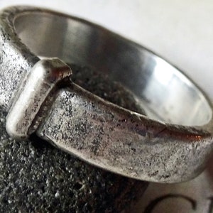 May include: A close-up of a silver ring with a textured, weathered appearance. The ring has a rectangular design element on the front. The ring is resting on a dark gray stone.
