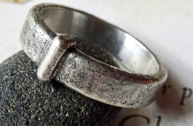 May include: A close-up of a silver ring with a textured, weathered appearance. The ring has a rectangular design element on the front. The ring is resting on a dark gray stone.