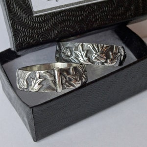 May include: Two silver rings with detailed floral and bird designs, presented in a black gift box. The rings feature a textured surface with intricate carvings of leaves, flowers, and birds. The box has a textured pattern.