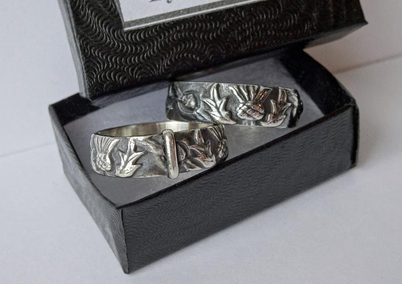 May include: Two silver rings with detailed floral and bird designs, presented in a black gift box. The rings feature a textured surface with intricate carvings of leaves, flowers, and birds. The box has a textured pattern.