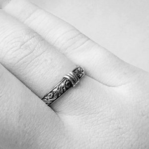 May include: A silver ring with an ornate floral design. The ring is worn on a finger, showcasing the intricate details of the metalwork. The ring's band features a detailed pattern, and the top has a wrapped design.