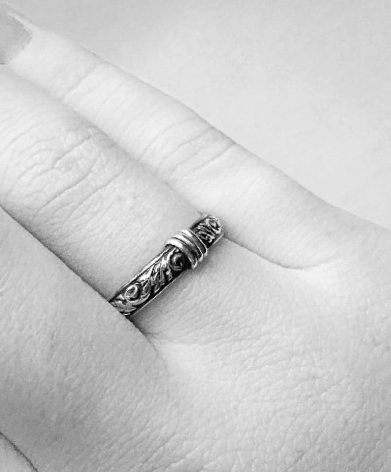 May include: A silver ring with an ornate floral design. The ring is worn on a finger, showcasing the intricate details of the metalwork. The ring's band features a detailed pattern, and the top has a wrapped design.