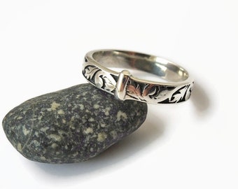 May include: A silver ring with an engraved floral pattern and a central bar detail. The ring is displayed on a dark gray stone against a white background. The ring's design features intricate leaf and vine motifs.