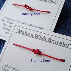 May include: Two red string bracelets with white cards. One card reads "Infinity Knot" and the other reads "Eternity Knot". The cards have text that says "Make a Wish Bracelet". The bracelets are on a denim background.