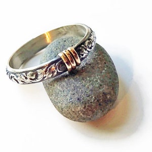 May include: A silver ring with an engraved floral pattern and gold accents. The ring is resting on a gray stone against a white background. The ring's design features intricate details and a contrasting color scheme.